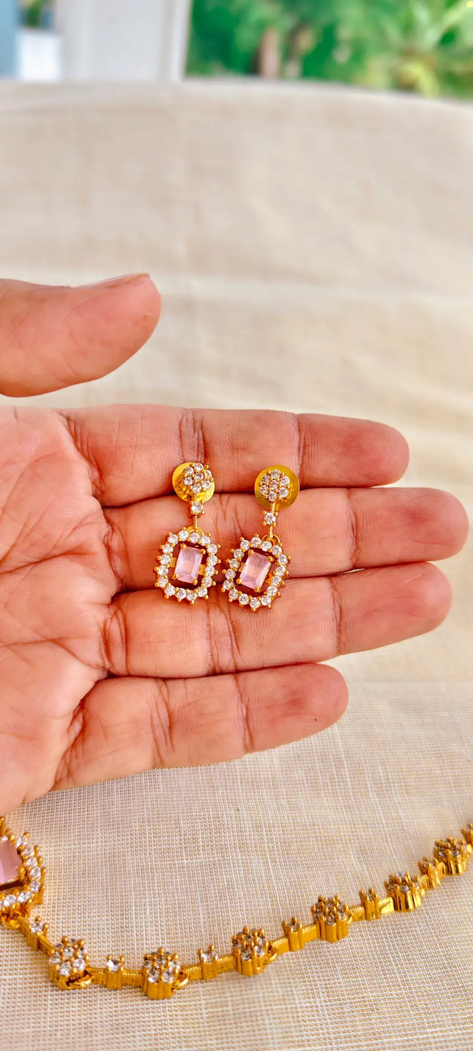 Gold earrings with pink gemstones held in a hand on a beige surface.