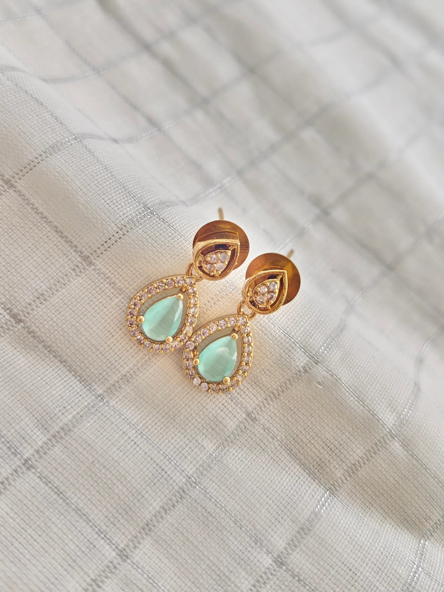 Gold earrings with green teardrop stones on a textured fabric background