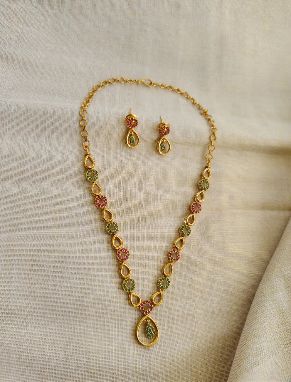 Gold necklace with green and red stones and matching earrings on a beige fabric background
